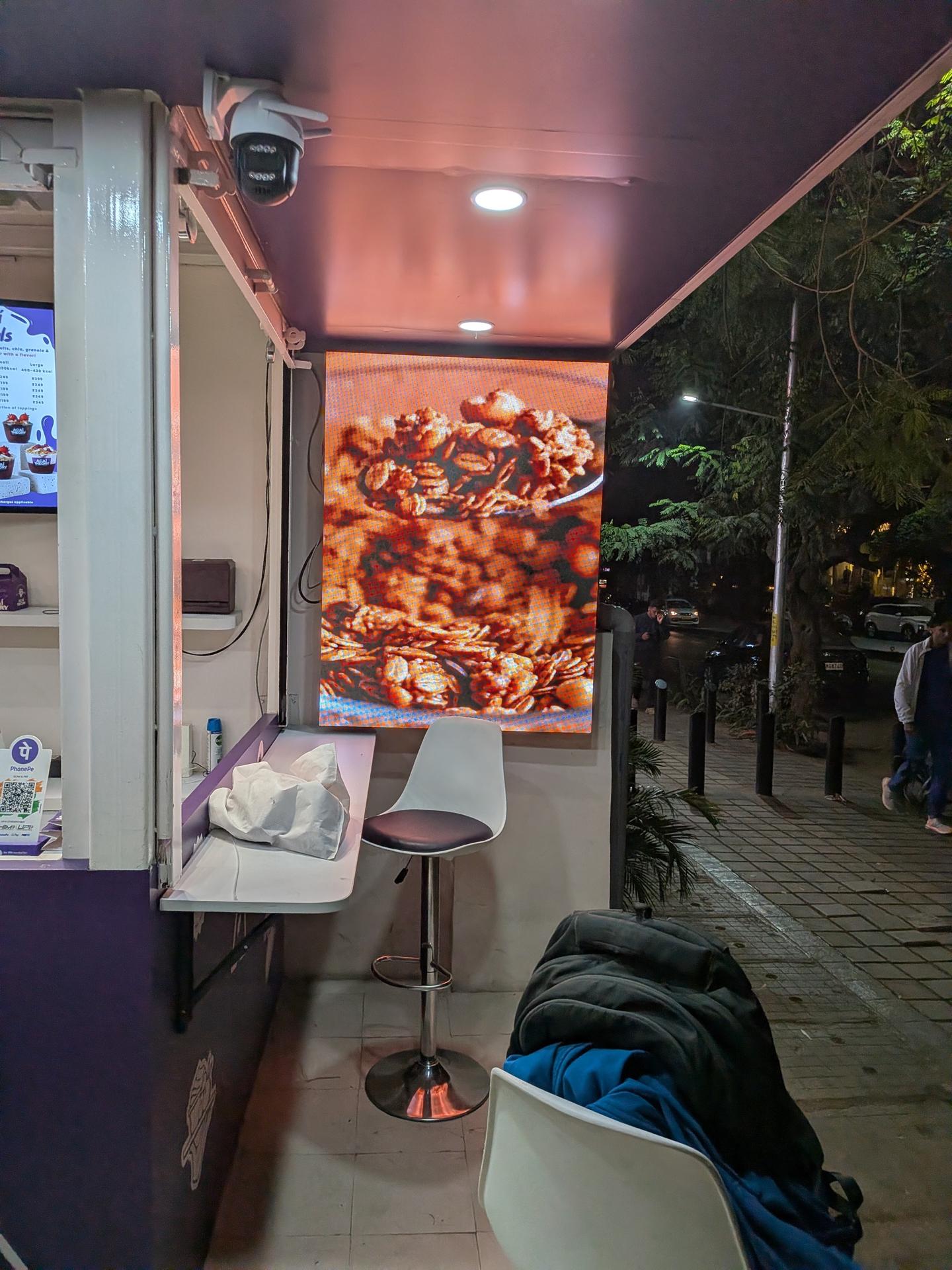 Portrait outdoor LED wall beside the counter of the Acai Theory shopfront in Indiranagar, Bengaluru, lit after dark and facing the pavement